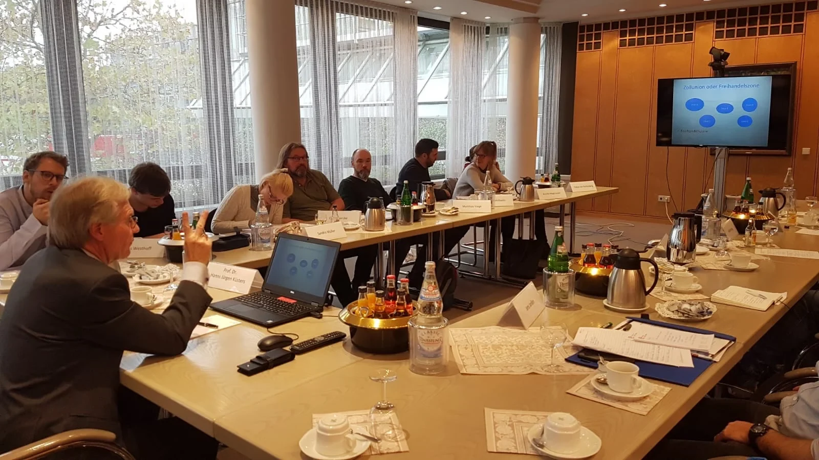 Eine Wissenschaftlerin und ein Wissenschaftler arbeiten hinter einer Glasfassade und mischen Chemikalien mit Großgeräten.