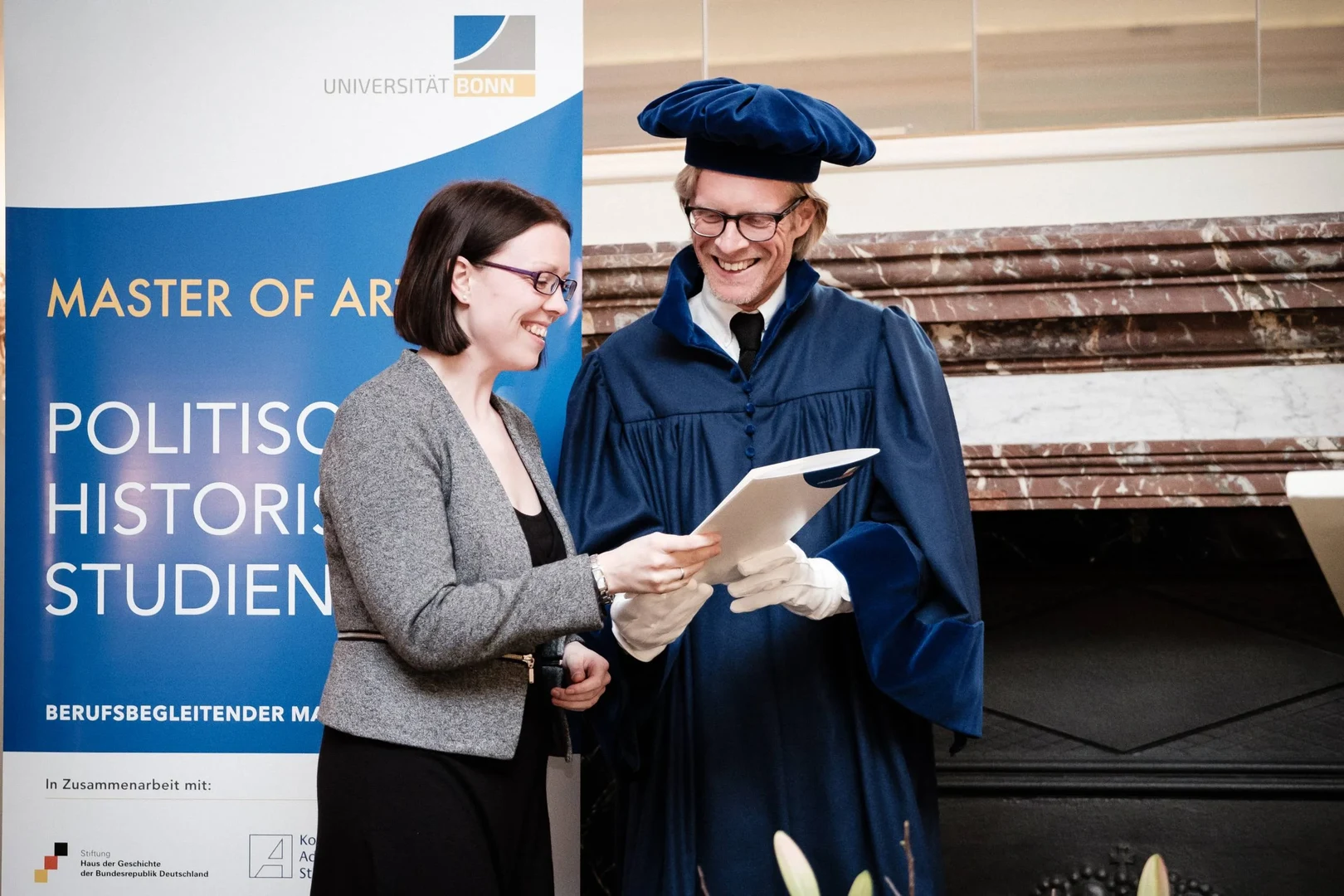 Eine Wissenschaftlerin und ein Wissenschaftler arbeiten hinter einer Glasfassade und mischen Chemikalien mit Großgeräten.